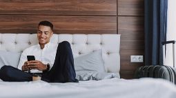 Cheerful man with smartphone is drinking coffee in hotel bedroom