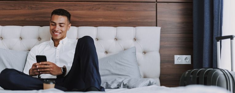 Allianz - Cheerful man with smartphone is drinking coffee in hotel bedroom